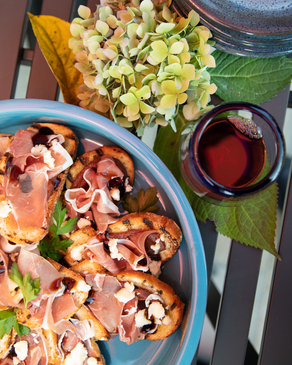 A colorful and appetizing spread of food, including grilled bruschetta topped with prosciutto, and a glass of red wine, set against a backdrop of vibrant yellow and green floral arrangements.