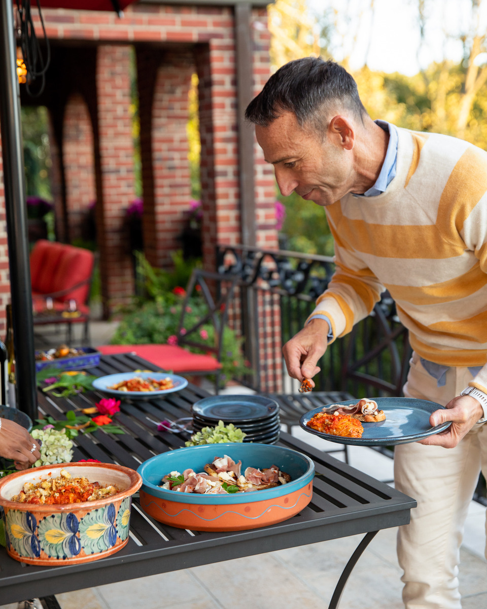 A man arranging and serving food on a patio table, surrounded by a variety of colorful dishes.