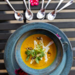 vibrant orange-colored soup served in a speckled blue-gray bowl, garnished with a dollop of coriander crème, crunchy pepita brittle, and fresh scallions and cilantro leaves.