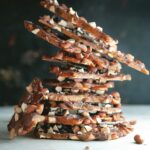 A stack of nut and chocolate salted brittle sits stacked on a white table with a dark background behind the subject.