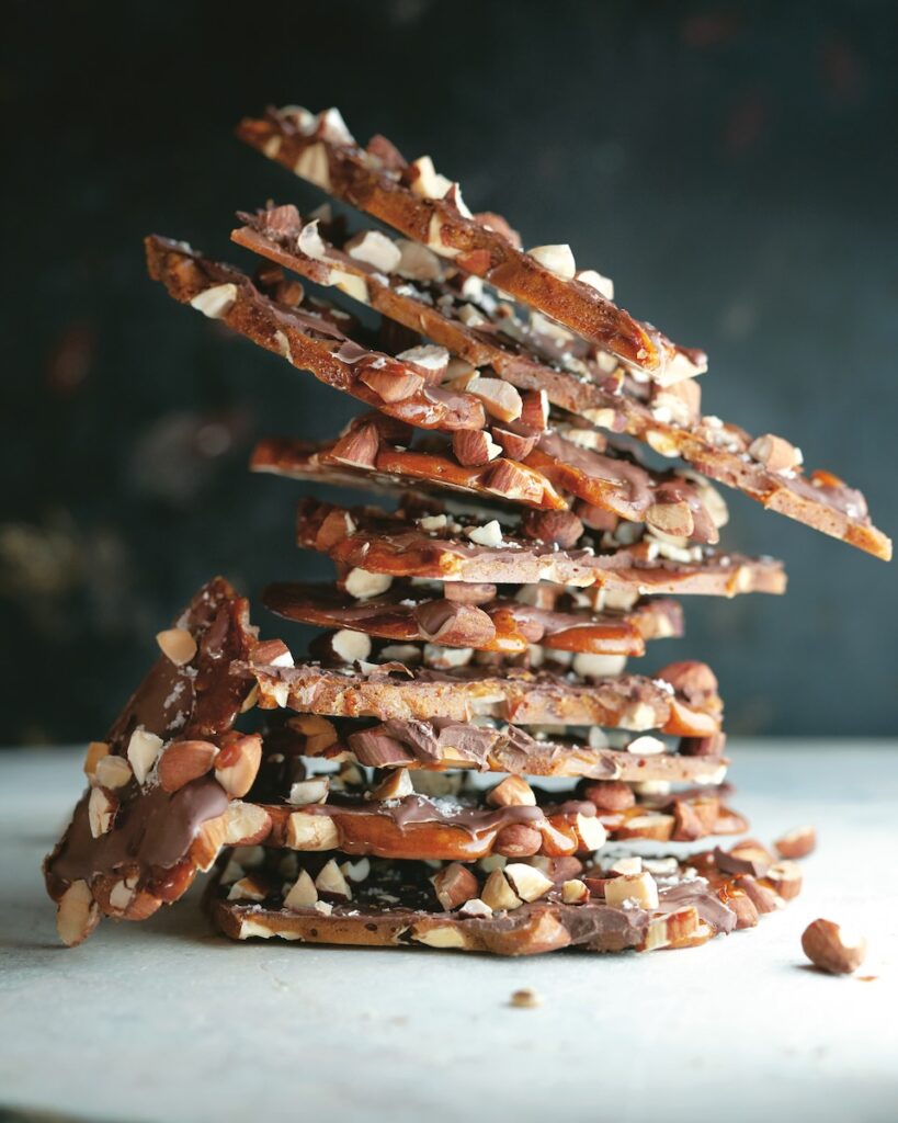 A stack of nut and chocolate salted brittle sits stacked on a white table with a dark background behind the subject.