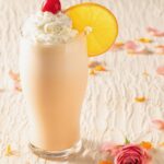 An orange creamsicle shake with whipped cream, a cherry on top, and an orange wheel garnish from Say Anything on a white table with rose petals.