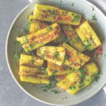 A large white bowl of smashed cucumbers with kimchi, parsley, and chili seasoning on top.