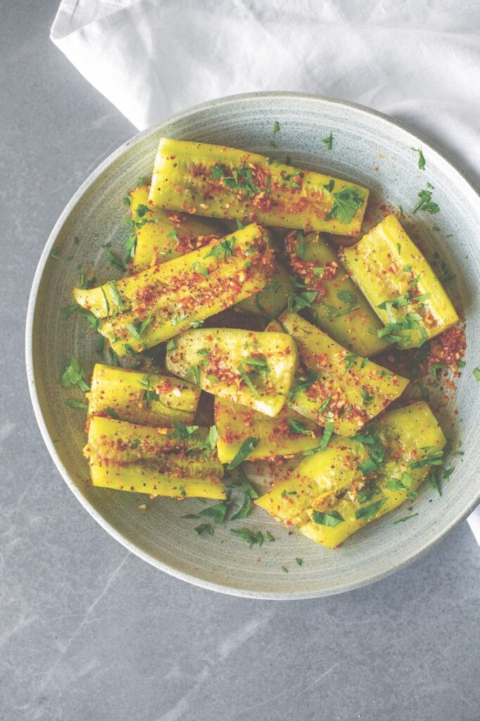 A large white bowl of smashed cucumbers with kimchi, parsley, and chili seasoning on top.