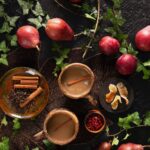 Two brown megs with coffee and a cinnamon stick inside sit next to a plate of cinnamon sticks and surrounded by red fruits on a green vine.