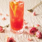 In a tall glass sits a red Sweet Home Alabama Slammer garnished with a heart formed orange peel and surrounded by pink roses on the table.