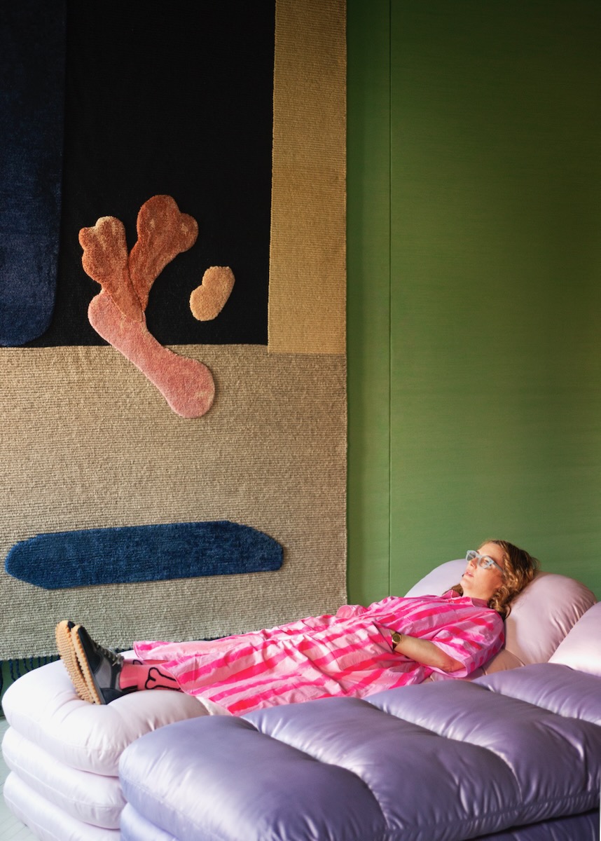 An interior by Faye Toogood with a green wall. A woman lays on a pink bed in a pink pajama set.