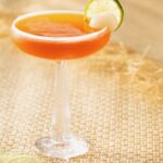 A tall martini glass holds an orangish thai getaway drink in honor of The White Lotus season with a lime wheel garnish.