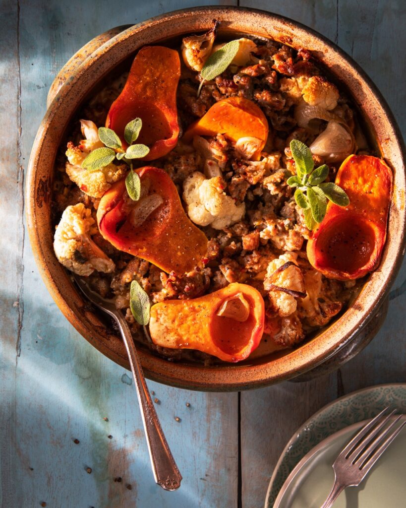 A bowl of Winter Veggie Bake with honeynut squash and cauliflower sits with a wood spoon inside it on a picnic table.