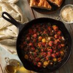 In a cast iron skiller sits a hot cherry tomato salad with cherry tomatoes and herbs as focaccia bread, cheese, and oil sit off to the side.