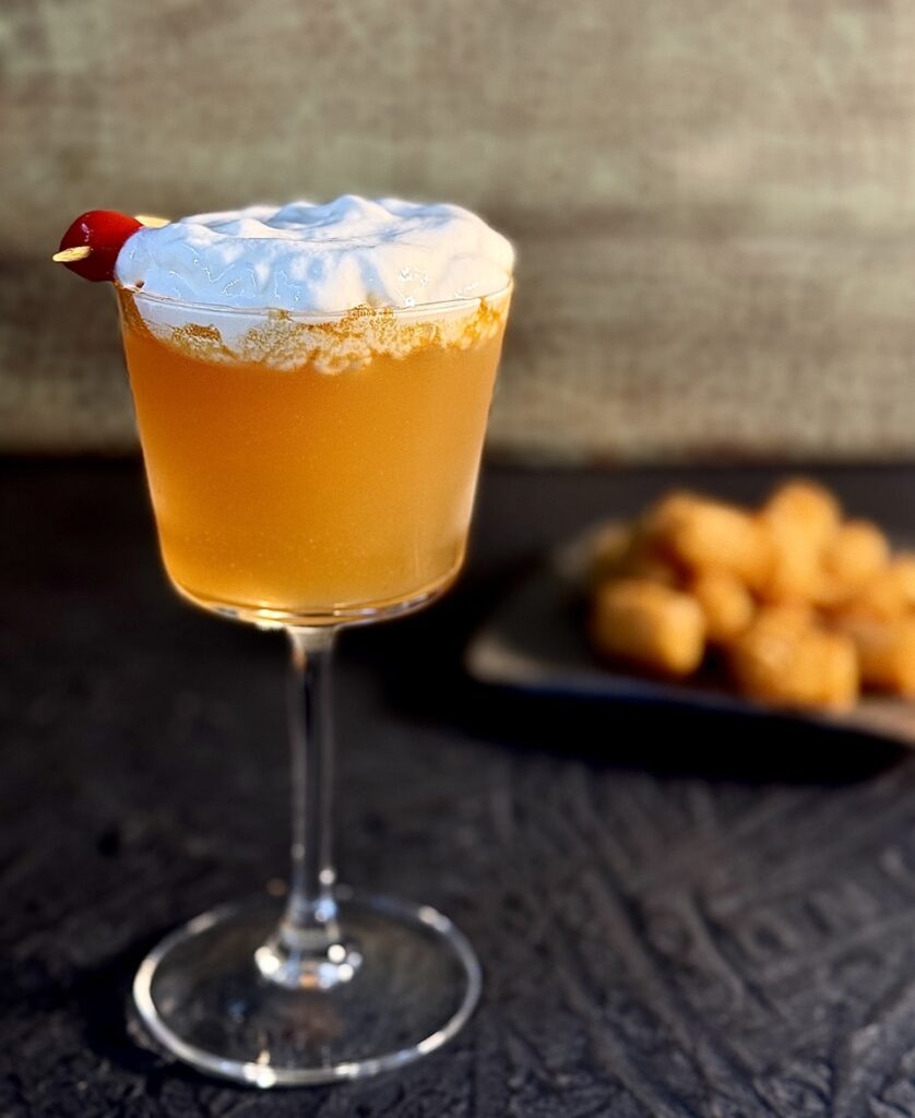 A tall cocktail glass holds a With Whiz cocktail for the Philadelphia Eagles with a foam on top and tater tots on a plate in the background.