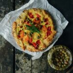 An Everything Bagel Tomato Tartelette sits on a piece of parchment paper with a jar of capers sitting nearby.