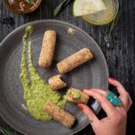 A woman with a ring on her finger picks up a log of Lamb Croquettes off of a grey plate with a green sauce smeared across it.