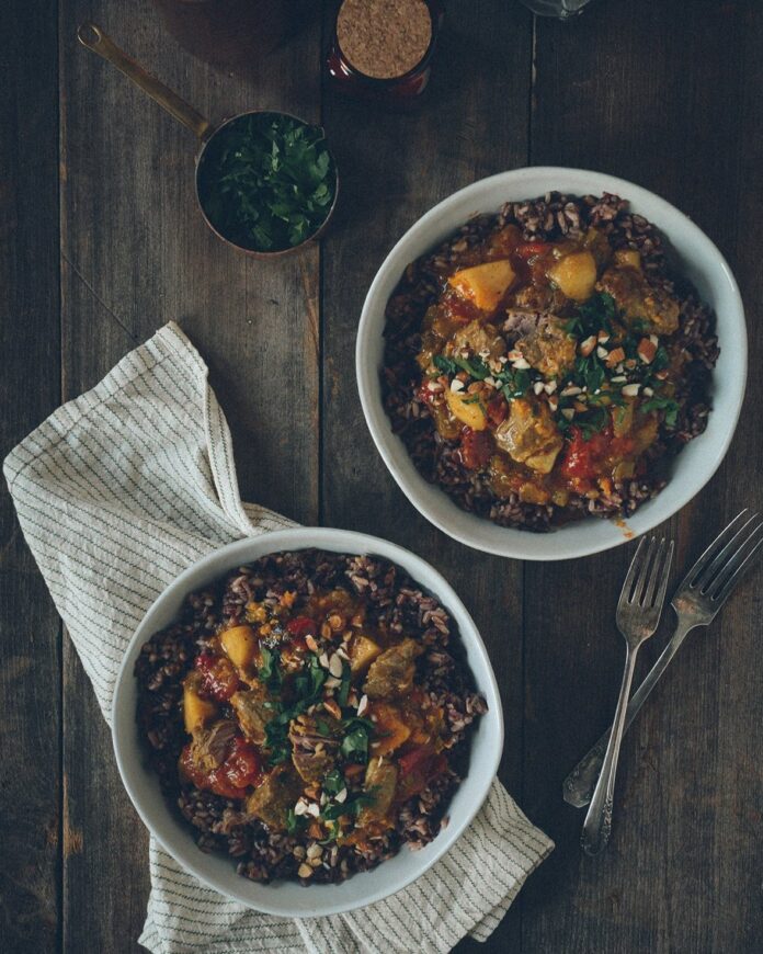 Two white bowls of a dark lamb curry loaded with vegetables on a dark picnic table as a white napkin sits underneath the one to the left.