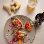 Two small hot dogs made out of lamb sausage sit with relish toppings on a grey plate as lamb donuts sit nearby.