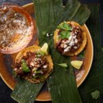 Two small tacos on green plantains hold a braised lamb shoulder with cilantro on top.