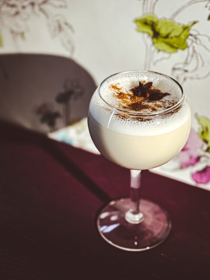 A round glass with a Jennifer Coolidge milk/milf punch cocktail inside a round glass topped with a nutmeg powder and star anise against a flower background.