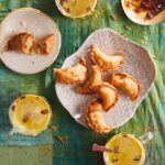 Six Sweet Gujiyas sit on a white plate as two more sit on another small plate. Three cups of hindu tea also sit scattered between the plates.