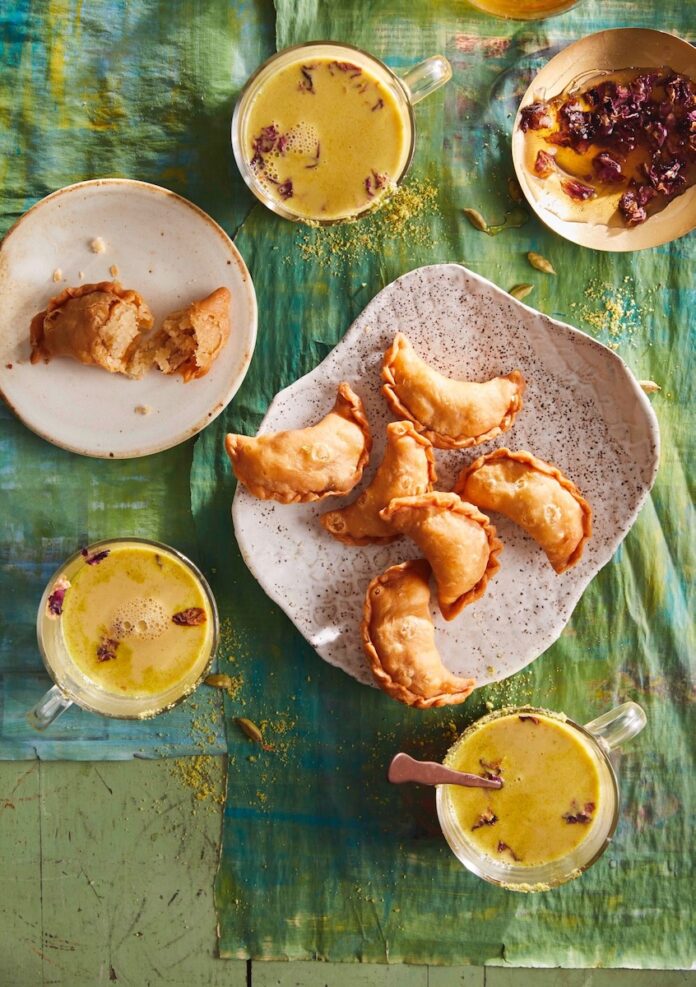 Six Sweet Gujiyas sit on a white plate as two more sit on another small plate. Three cups of hindu tea also sit scattered between the plates.